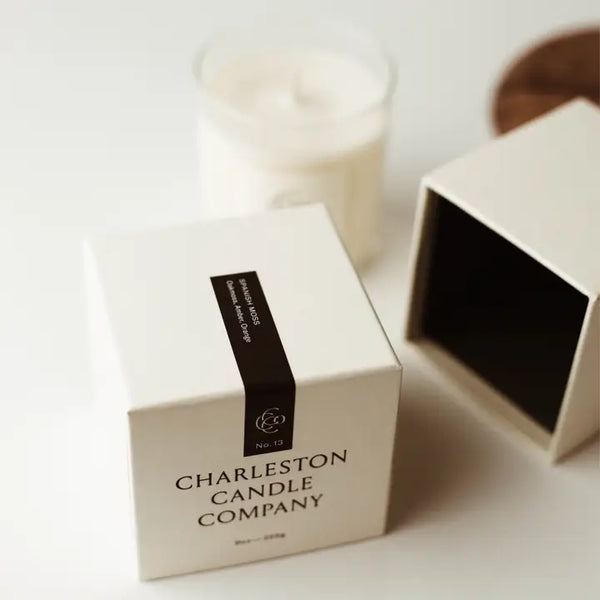 A white Charleston Candle Company box with a dark label strip next to an open box and a lit white candle in the background, evoking a cozy, relaxing ambiance.