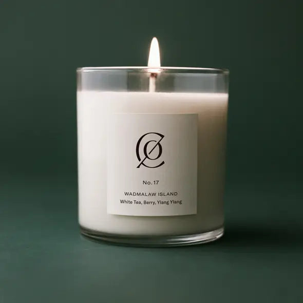 Lit white candle in a clear glass holder labeled with No. 17 Wadmalaw Island, featuring scents of white tea, berry, and ylang ylang against a dark green background.
