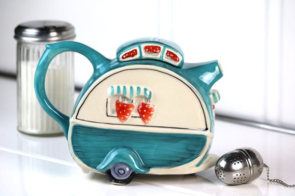 Ceramic teapot shaped like a vintage dining car in teal and cream colors with red accents, displayed next to a metal tea infuser and sugar container.