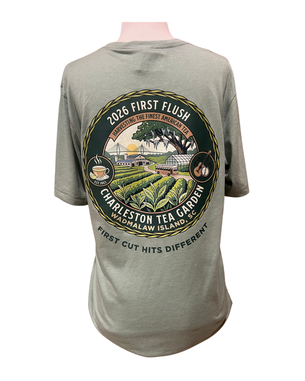 Back of a soft green t-shirt featuring a colorful Charleston Tea Garden design, showcasing tea fields, a tea cup, and a guitar in a circular layout celebrating the 2026 first flush season.