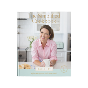 A smiling woman in a pink shirt holding a coffee mug, sitting in a bright modern kitchen with baked treats on the counter, suggesting easy and delicious home cooking.