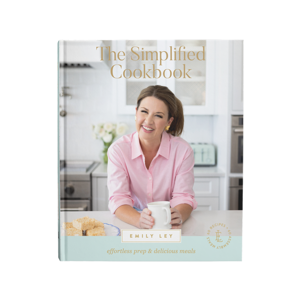 A smiling woman in a pink shirt holding a coffee mug, sitting in a bright modern kitchen with baked treats on the counter, suggesting easy and delicious home cooking.
