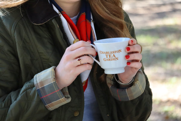 Person holding a white ceramic cup with "Charleston Tea Garden" logo, dressed in a green jacket with plaid cuffs and a red scarf outdoors.