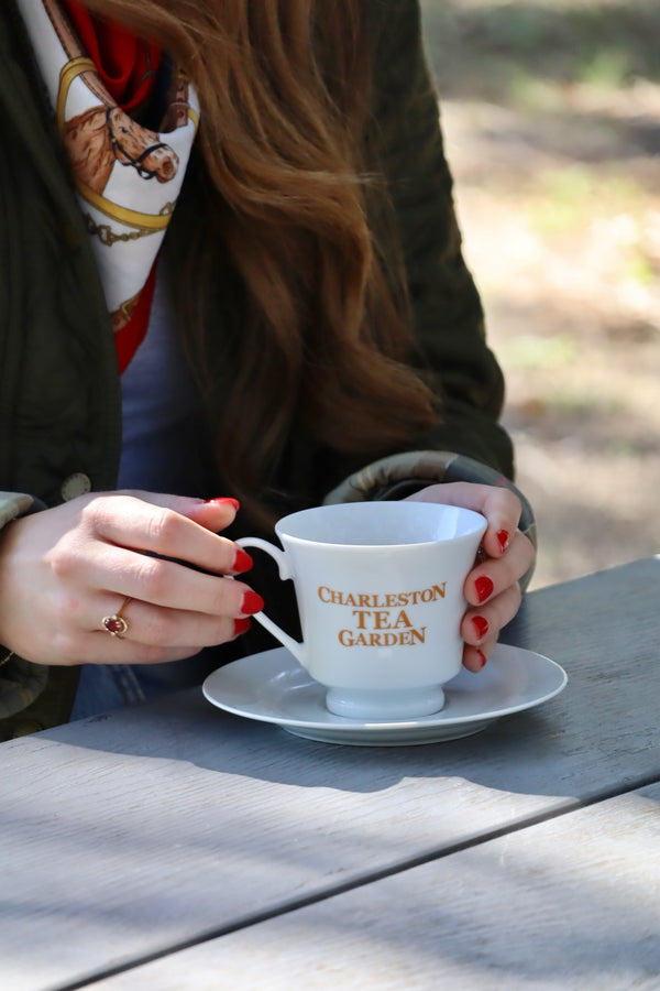 Hands with red nail polish holding a white tea cup and saucer with gold lettering that reads Charleston Tea Garden, set on a wooden table outdoors.