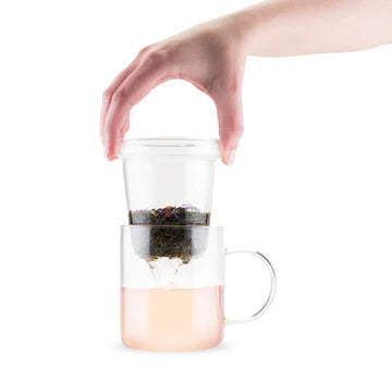 Hand placing a glass tea infuser filled with loose leaf tea into a clear glass mug with light pink brewed tea, showcasing a simple and elegant brewing process.