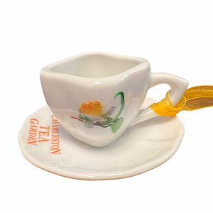 Small white ceramic tea cup ornament with matching saucer, decorated with a delicate floral design and "Charleston Tea Garden" text, featuring a yellow ribbon for hanging.