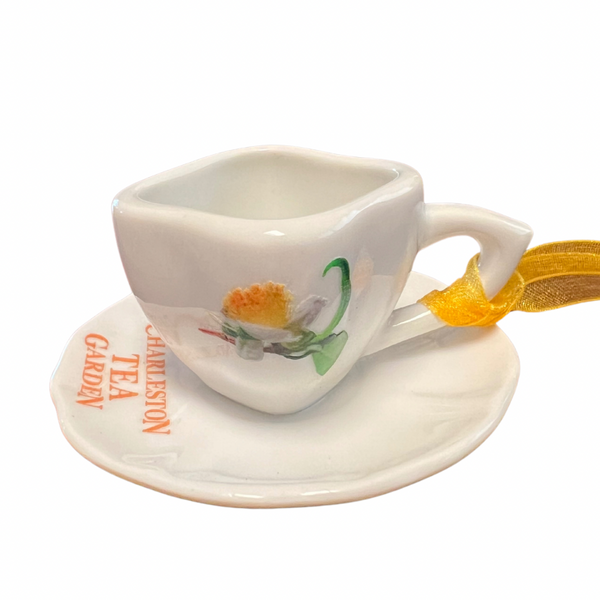 Small white ceramic tea cup ornament with matching saucer, decorated with a delicate floral design and "Charleston Tea Garden" text, featuring a yellow ribbon for hanging.