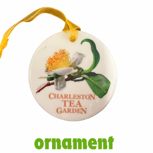 Round ceramic ornament with a painted tea flower and green leaves, featuring the words Charleston Tea Garden, hanging by a yellow ribbon.