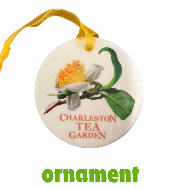 Round ceramic ornament with a painted tea flower and green leaves, featuring the words Charleston Tea Garden, hanging by a yellow ribbon.