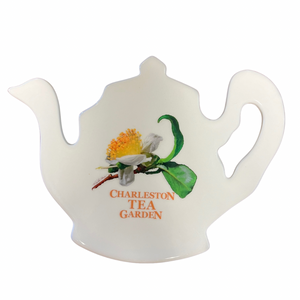 White ceramic tea pot-shaped trivet featuring a delicate illustration of a tea flower and green leaves with “Charleston Tea Garden” printed in orange.