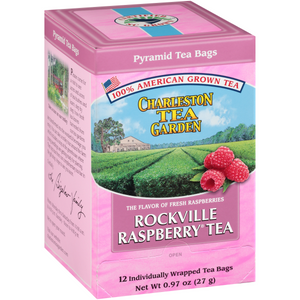 Pink box of Charleston Tea Garden pyramid tea bags featuring fresh raspberries and a scenic view of lush green tea fields under a blue sky.