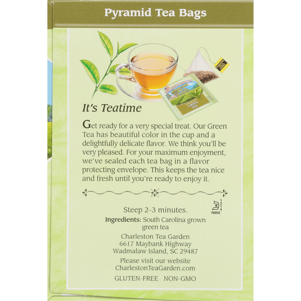 Close-up of pyramid-shaped green tea bags next to a clear glass cup filled with light green tea, accompanied by fresh green tea leaves and soft sunlight on a pale green background.