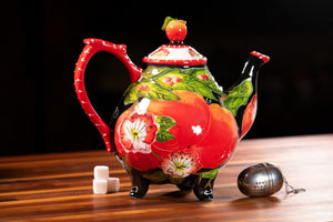 Colorful teapot with a red handle and vibrant apple and floral designs, sitting on a wooden surface with sugar cubes and a tea infuser nearby.