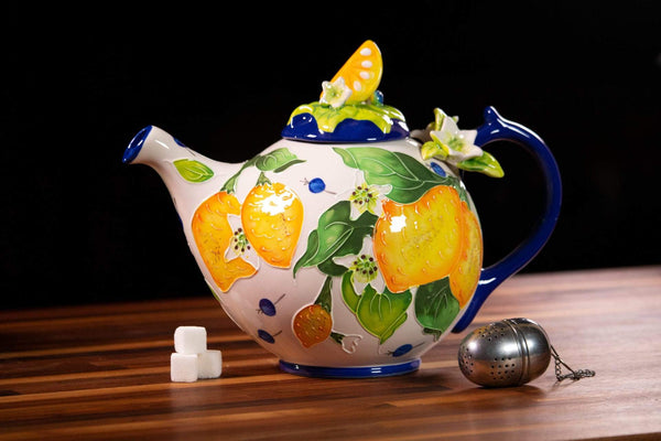 Colorful ceramic teapot decorated with bright yellow lemons and green leaves, accompanied by a metal tea infuser and sugar cubes on a wooden surface.