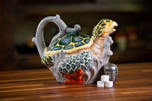 Ceramic teapot shaped like a sea turtle with detailed green and yellow shell design, sitting on a wooden surface next to sugar cubes and a metal tea infuser.